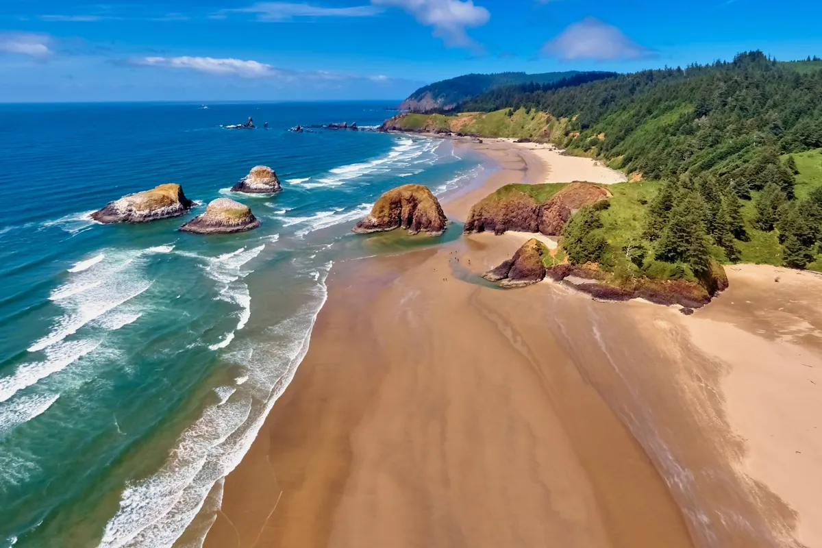 A stunning coastal scene featuring sandy beach, unique rock formations, gentle waves, and lush green hills in the background. The mood is peaceful, inviting families to beachcomb, hike, picnic, and explore tide pools at sunset.