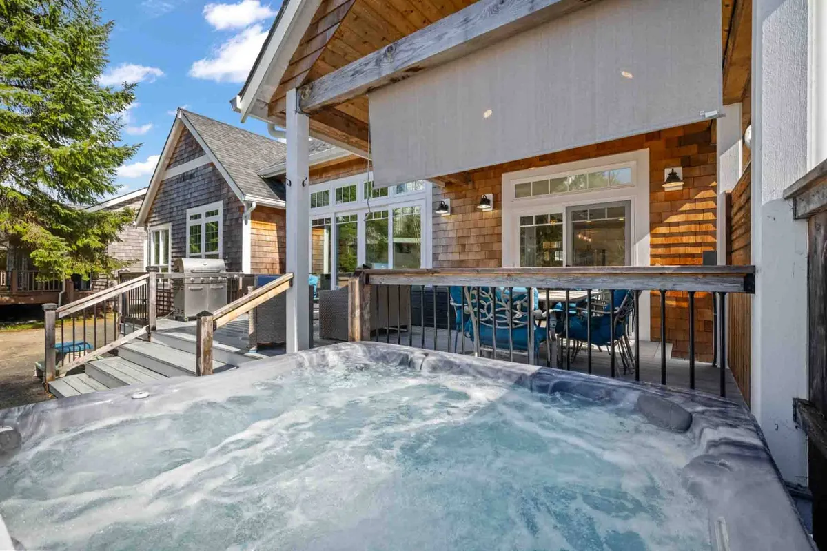 This image captures a spacious wooden deck surrounded by lush trees and features a private hot tub, an inviting outdoor dining area, and comfortable seating. The scene is set for relaxation and social gatherings, with natural light filtering through the foliage and hints of sunset visible in the background.