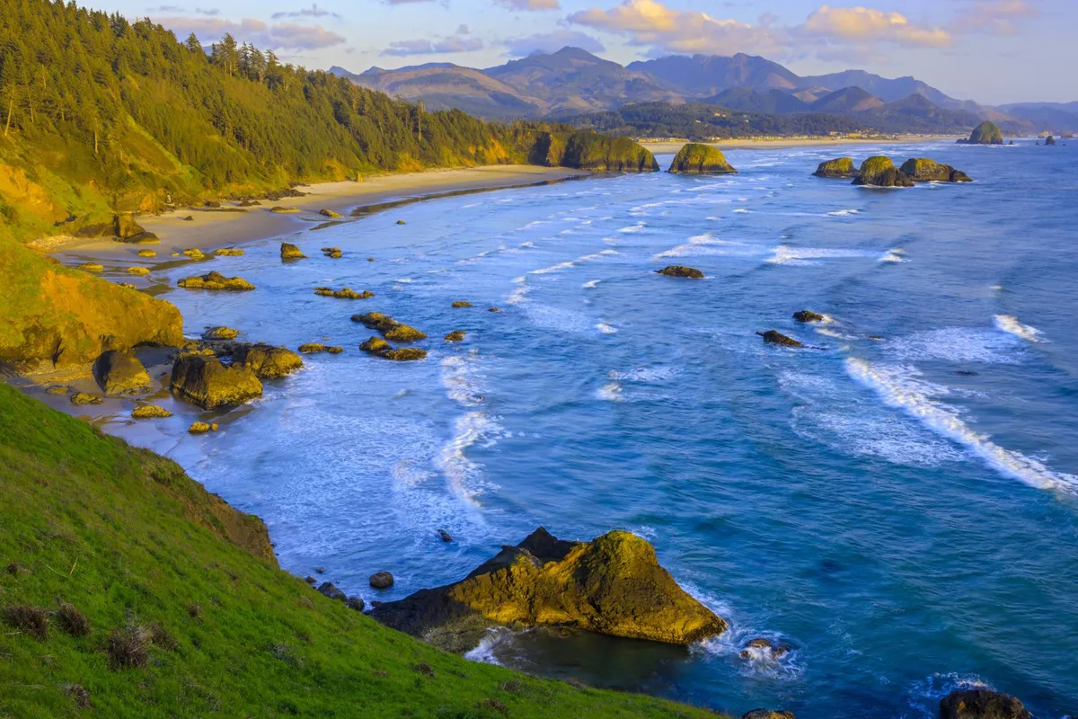 A stunning view of waves crashing against rocky outcrops, with lush green hills and distant mountains framing a calm, sandy beach. The expansive ocean and golden sunlight set a tranquil mood, inviting visitors to explore the natural beauty of the Oregon Coast.
