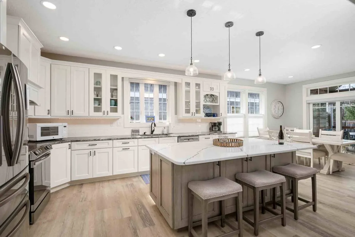 A well-lit, modern kitchen with a prominent island, stylish hanging pendant lights, and large windows. The space is designed for both cooking and socializing, with ample room for preparing meals and gathering around the island for casual dining or celebration.