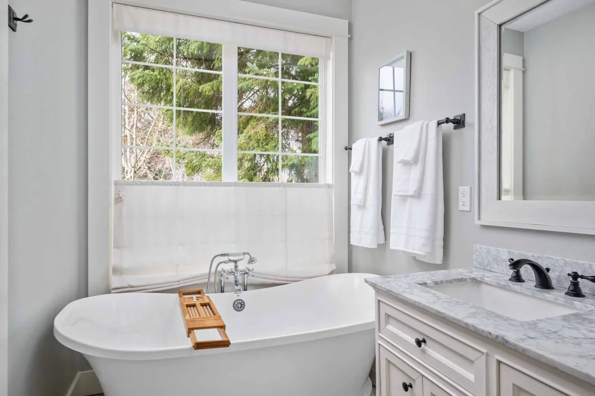 A spa-inspired bathroom with a freestanding bathtub positioned beneath a large window, marble countertop, and fluffy white towels. The space is filled with natural light, creating a soothing environment for self-care and relaxation.