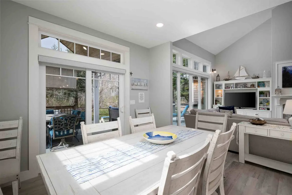 A bright, welcoming interior featuring a spacious dining table set near expansive windows that fill the open-plan living and dining area with natural light. The room includes a cozy living area with plush seating, decorative shelving with curated coastal décor, and an inviting ambiance ideal for social gatherings or relaxing family meals.