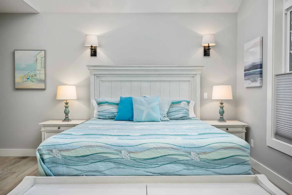 A tranquil bedroom featuring a bed dressed in aqua and white bedding, flanked by nightstands with elegant lamps and adorned with serene wall art. The soft natural light streaming in enhances the calming atmosphere, ideal for rest and relaxation.