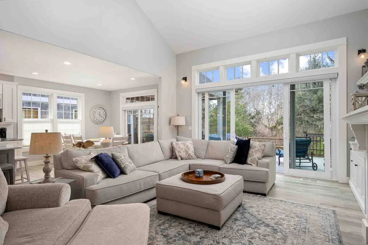 The image showcases a spacious, sunlit living room with a large plush sectional sofa adorned with decorative pillows, a stylish coffee table, and inviting sliding glass doors that open to serene views. The ambiance is cozy and bright, ideal for gatherings and relaxation.
